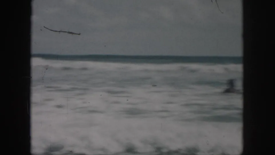 1958 8mm frame showing a blurred swimmer low in the surf, with choppy water and visible film wear.