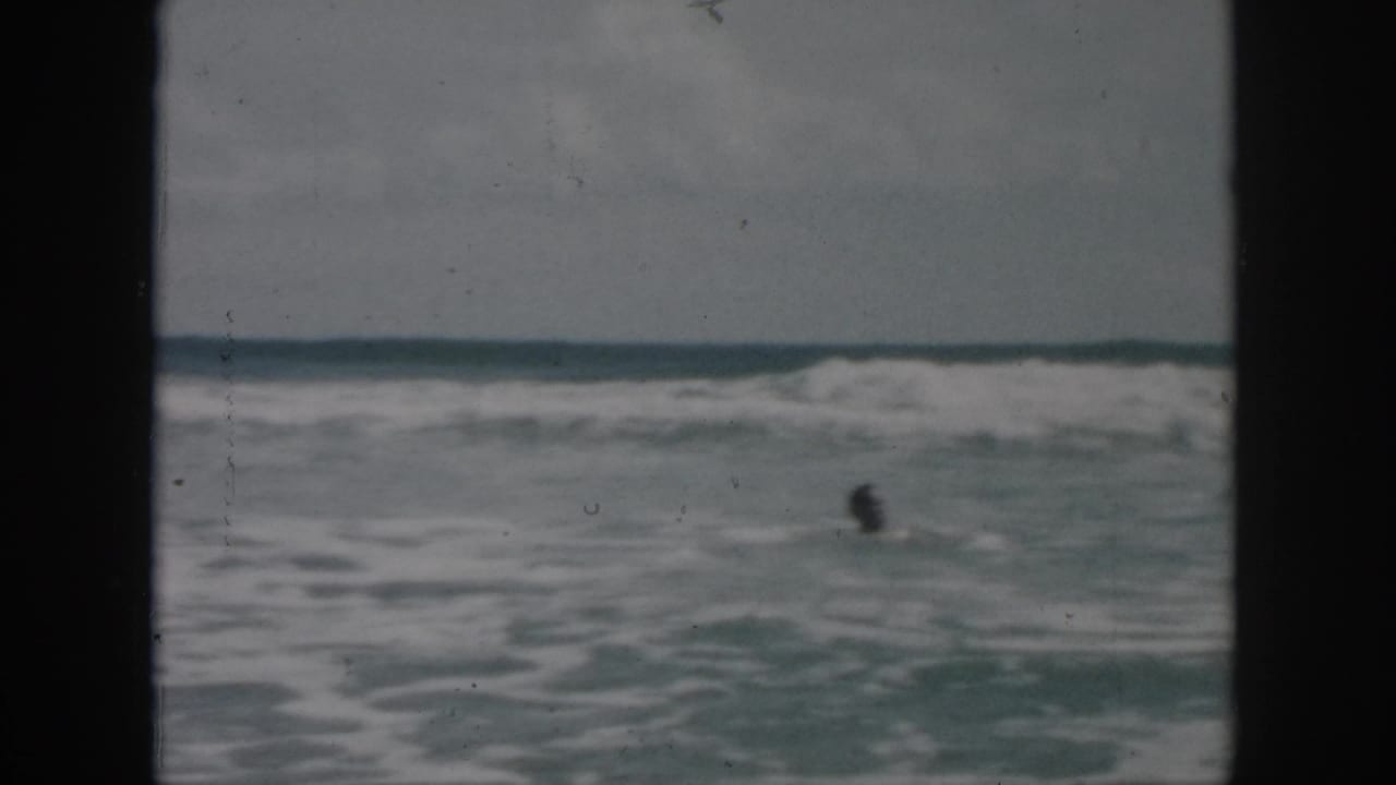 1958 8mm frame of a distant man swimming in rough ocean water near New Orleans, with breaking waves and a low horizon.