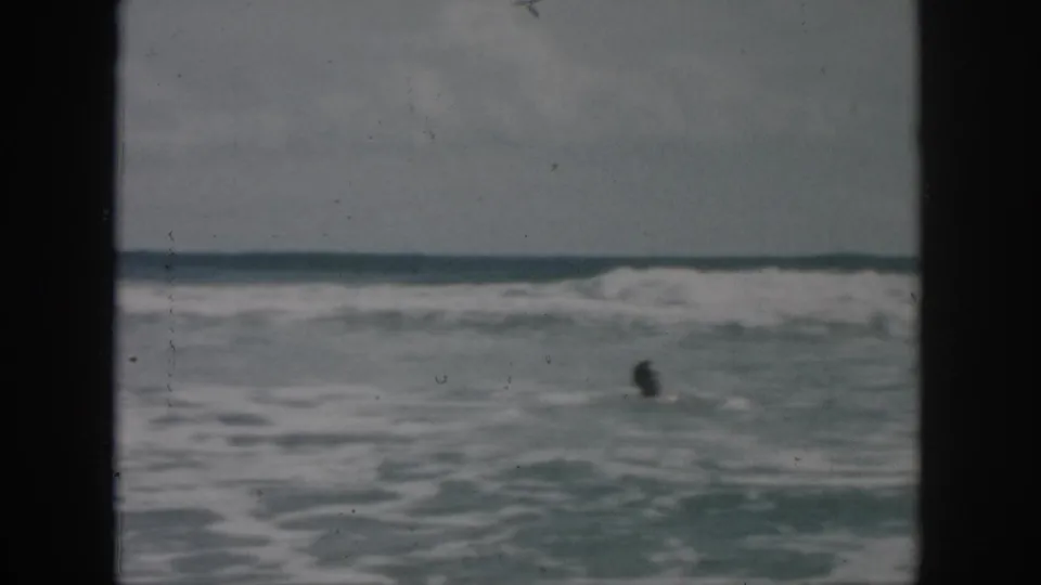 1958 8mm frame of a distant man swimming in rough ocean water near New Orleans, with breaking waves and a low horizon.