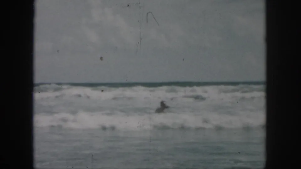 1958 8mm frame of a swimmer partly lost in choppy surf, with pale sky and ocean horizon visible.