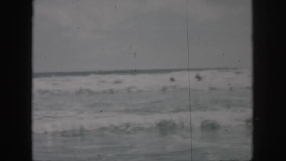 1958 8mm home-movie frame of rough surf near New Orleans, with soft focus, gray sky, and dark film gate edges.