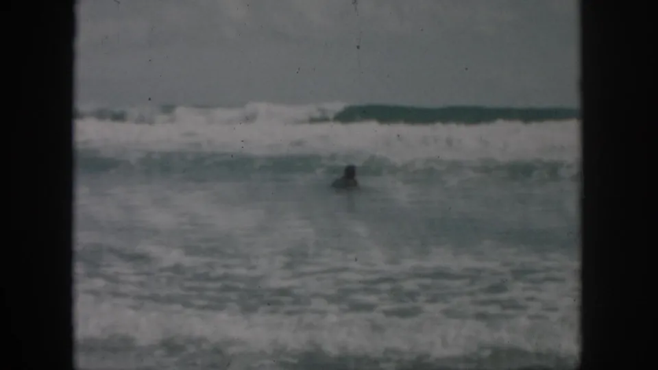Distant swimmer in choppy coastal water, filmed in soft 1958 8mm home-movie texture with breaking waves behind.