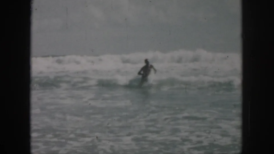 1958 8mm frame of an unidentified swimmer wading through rough surf near New Orleans, with muted color and visible film scratches.