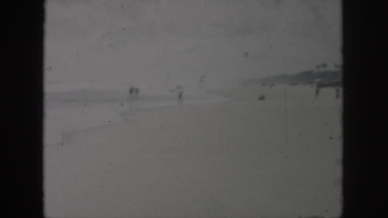 1958 New Orleans-area 8mm frame showing a low-contrast beach with distant people, shoreline haze, and faint pier-like structures.