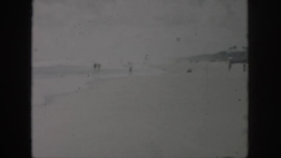 1958 New Orleans-area 8mm frame showing a low-contrast beach with distant people, shoreline haze, and faint pier-like structures.