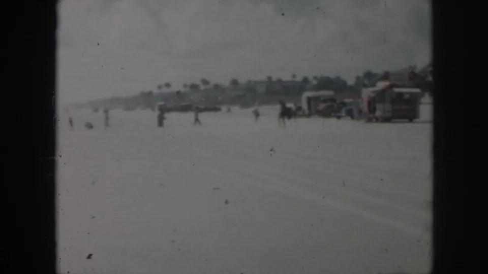 1958 New Orleans-area home-movie frame showing a beachside road or open sand area with people, buildings, and parked or moving cars.