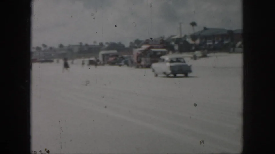 1958 New Orleans-area 8mm frame with a light-colored classic car passing beachside structures and small distant pedestrians.