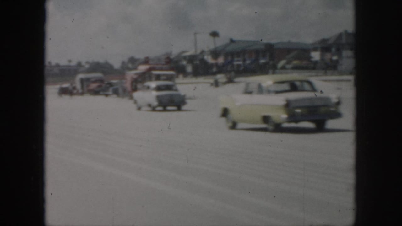 1958 New Orleans home-movie frame showing mid-century cars moving along a broad road beside low-rise buildings.