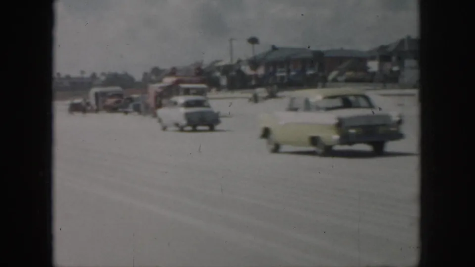 1958 New Orleans home-movie frame showing mid-century cars moving along a broad road beside low-rise buildings.
