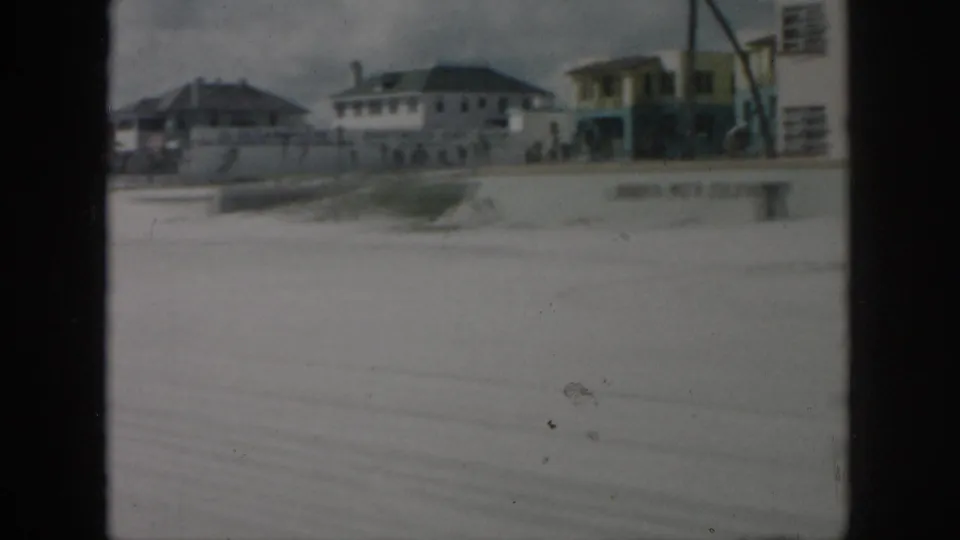 1958 New Orleans street-side view with low buildings, a painted wall, and small distant pedestrians across the road.