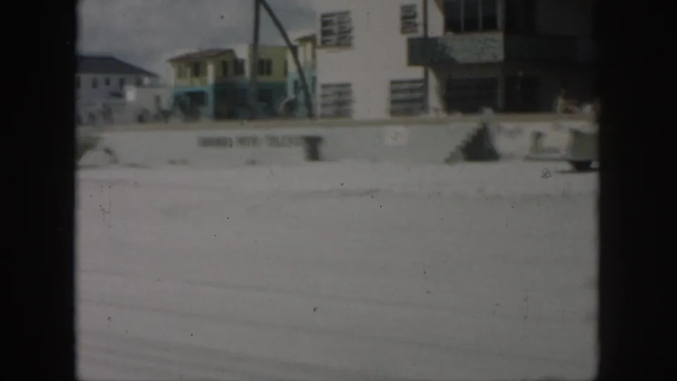 New Orleans 1958 home-movie frame with a partial car, low buildings, wall lettering, and small figures in the distance.