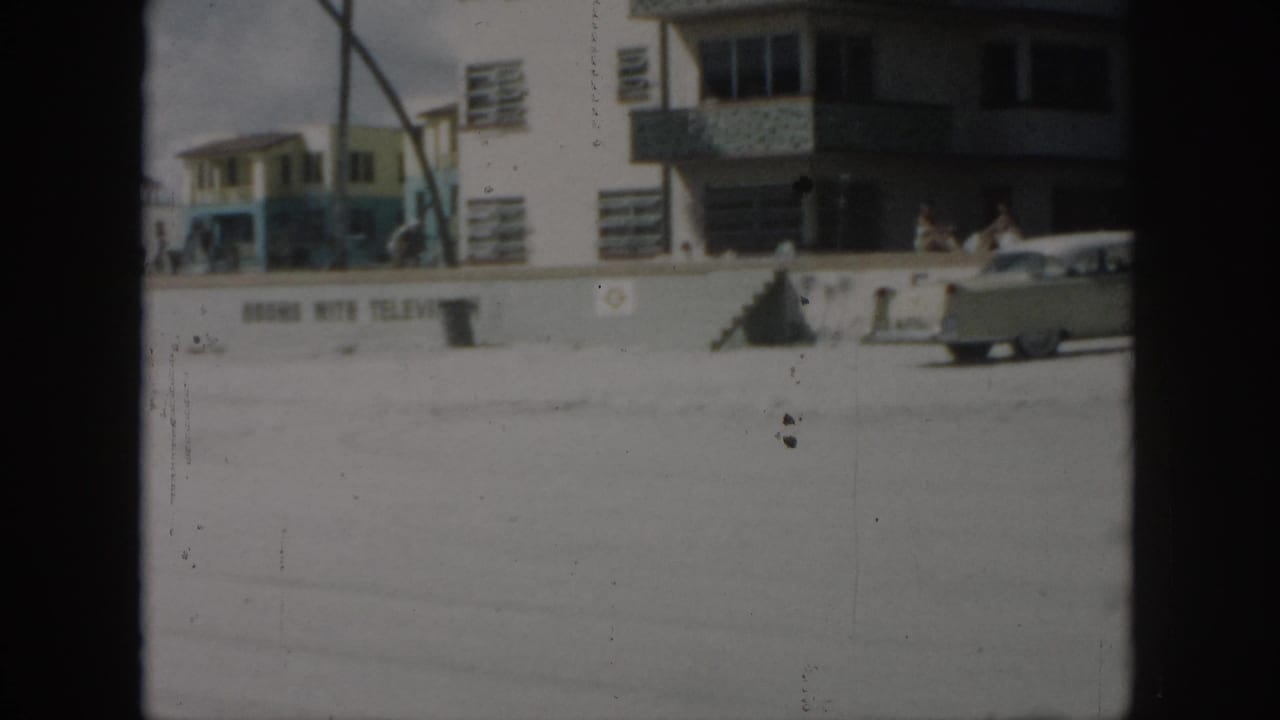 Light-colored sedan near a beachfront building with people along a seawall.