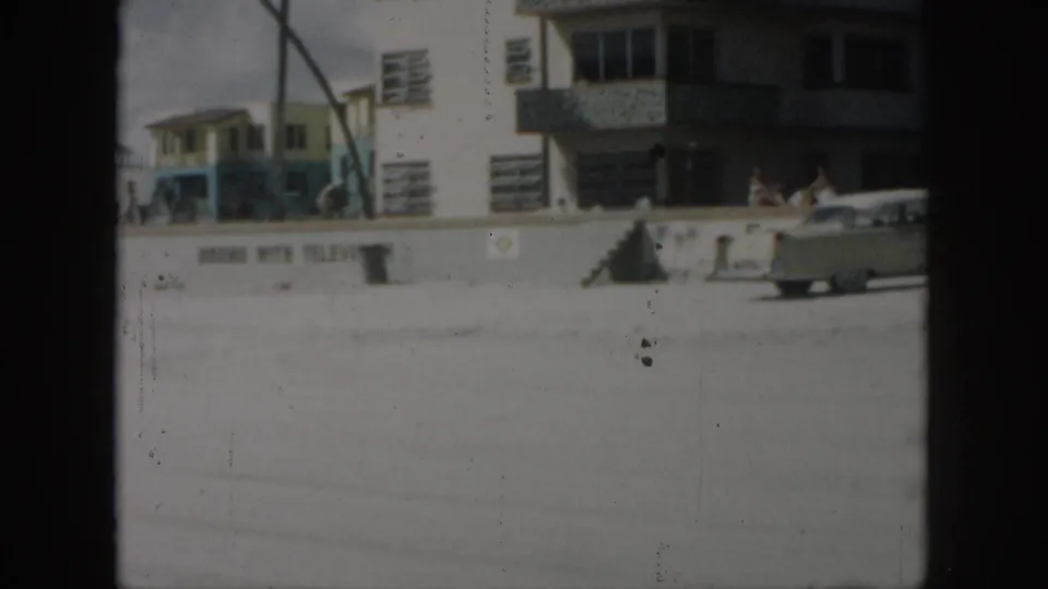 1958 New Orleans home-movie frame showing a pale sedan by a beach wall, a multi-story building, and small figures near the shore.