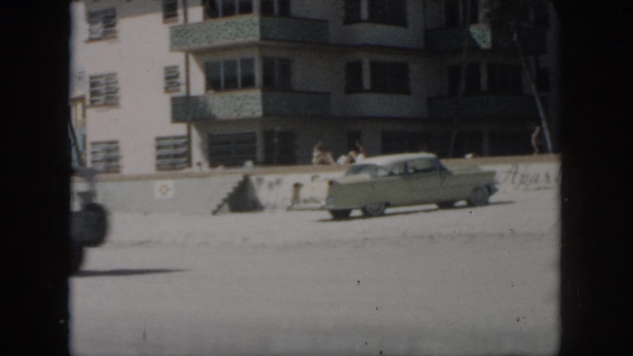 Pale sedan crossing in front of a beachfront building and seawall.