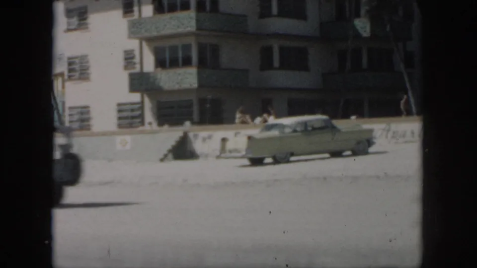 1958 New Orleans frame with a pale sedan moving across the foreground, people near the wall, and a beachside building behind them.