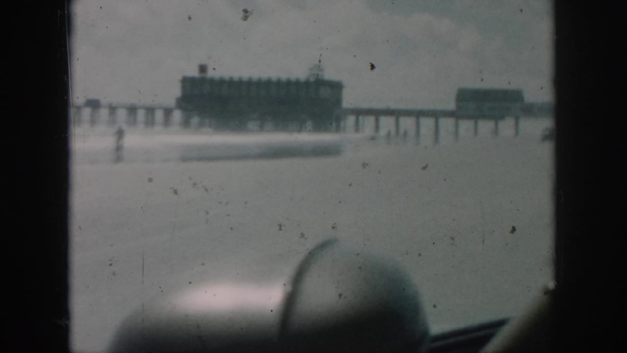 A 1958 home-movie frame from New Orleans showing pier structures beyond the beach, filmed through a vehicle with muted color and dust marks.