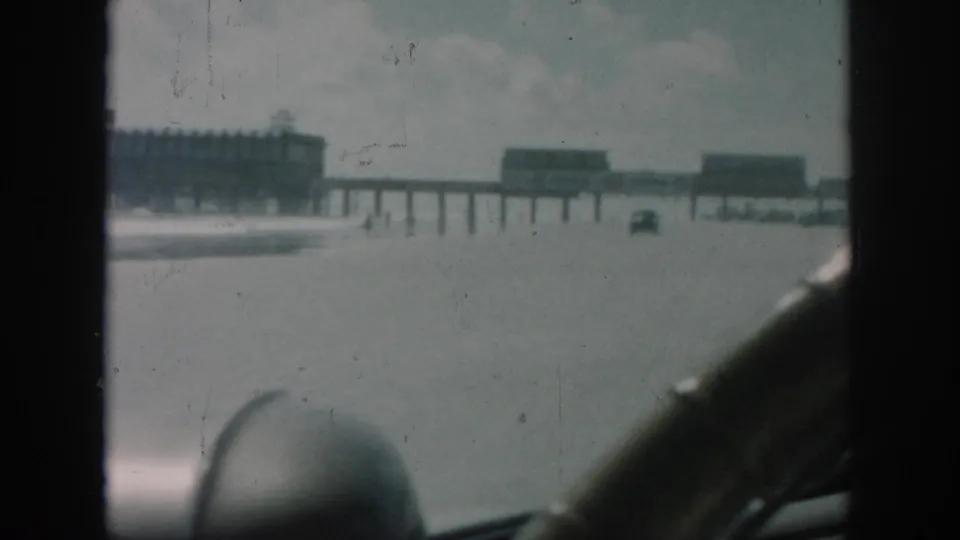 1958 8mm travel view of a New Orleans pier area, with distant structures, a small vehicle, and the car interior framing the lower foreground.