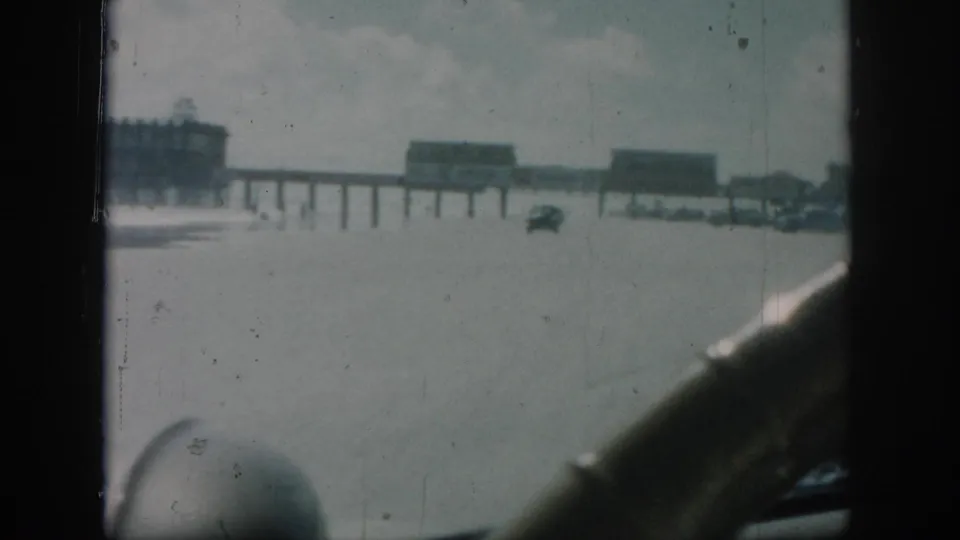 New Orleans 1958 home-movie frame with pier buildings, a small distant vehicle, and dark car interior shapes at the right and lower edges.