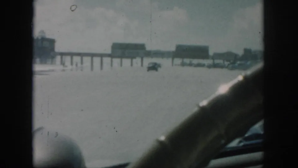 1958 New Orleans home-movie view from inside a car approaching a flooded road with distant cars and elevated structures.