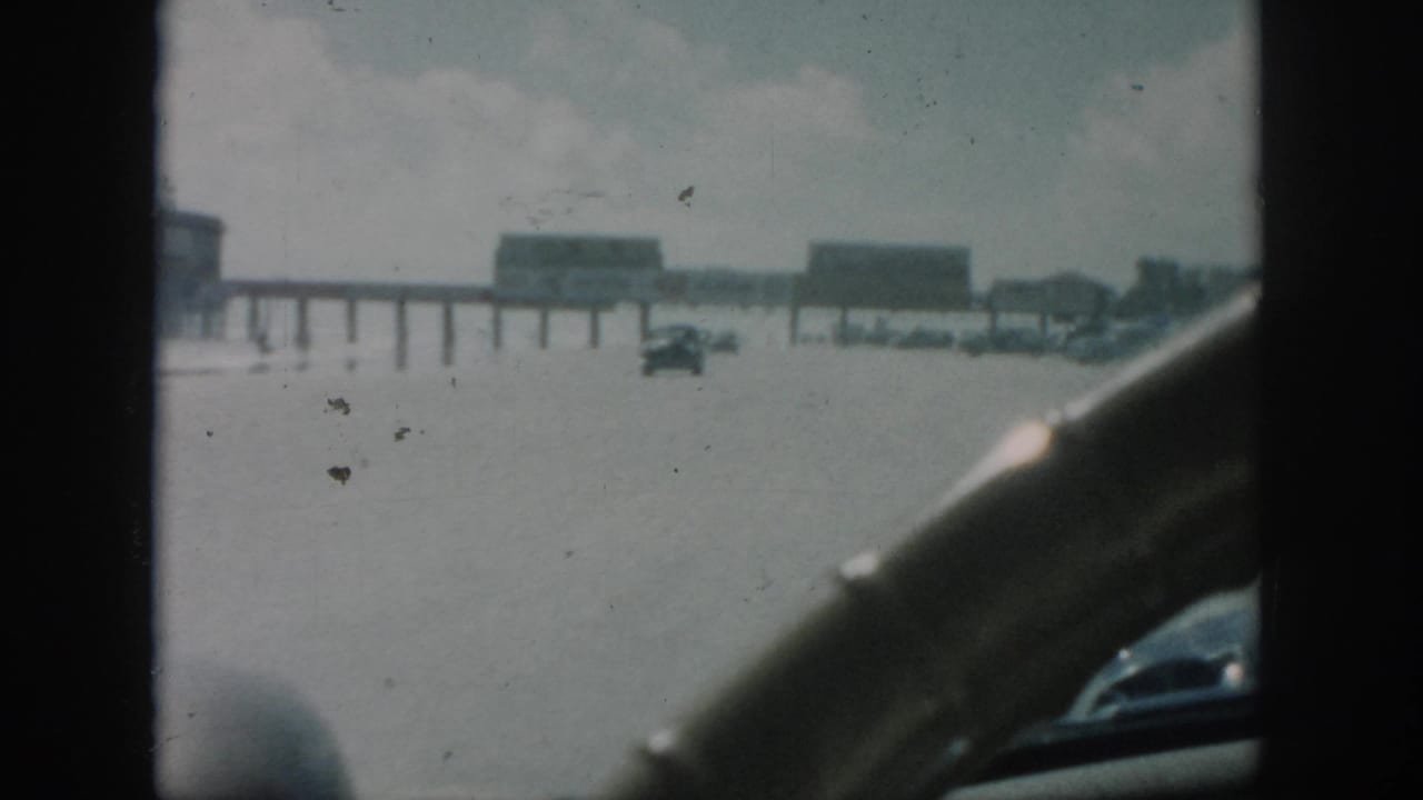 Flooded road seen through a car window with a vehicle ahead and elevated structures in the distance.