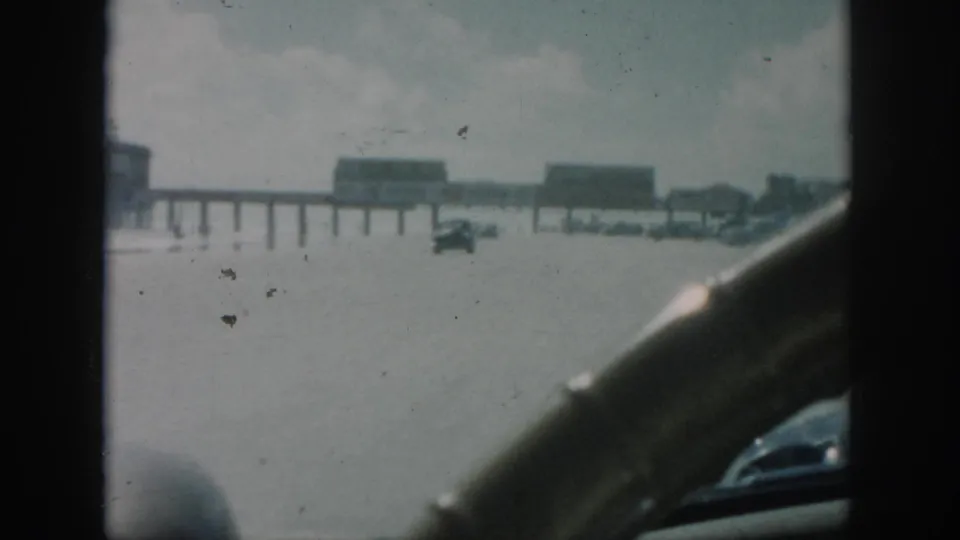 Inside-car view in 1958 New Orleans showing high water over the road, a car ahead, and distant pier-like buildings.