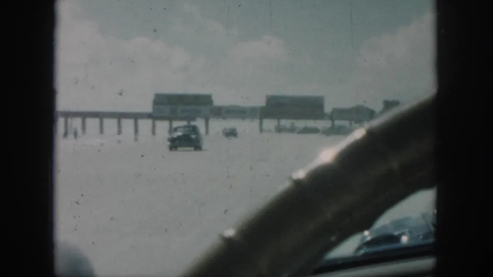 Home-movie frame from 1958 New Orleans showing cars crossing a flooded roadway beyond the windshield and dashboard.