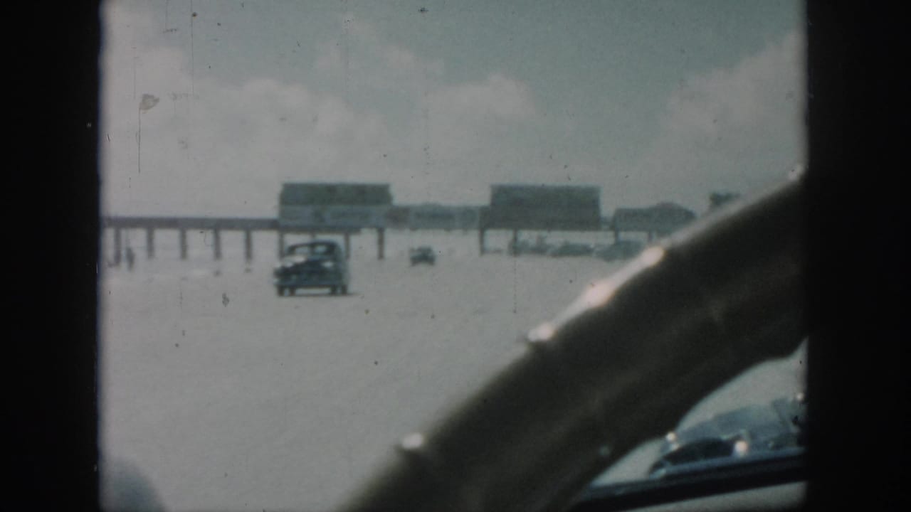 1958 New Orleans home-movie view from inside a moving car, with a steering wheel framing a road, distant cars and a long pier-like structure.