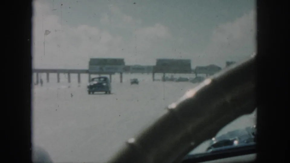 1958 New Orleans home-movie view from inside a moving car, with a steering wheel framing a road, distant cars and a long pier-like structure.