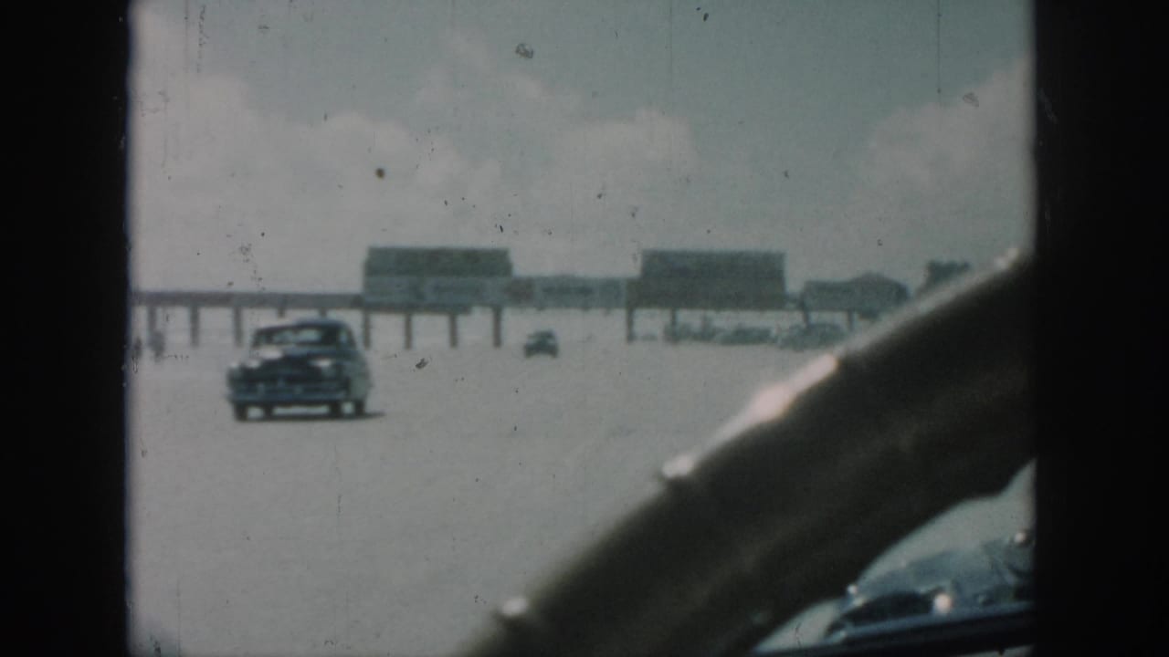 Windshield view of a broad road, distant cars and a long structure on posts, partly framed by a steering wheel.