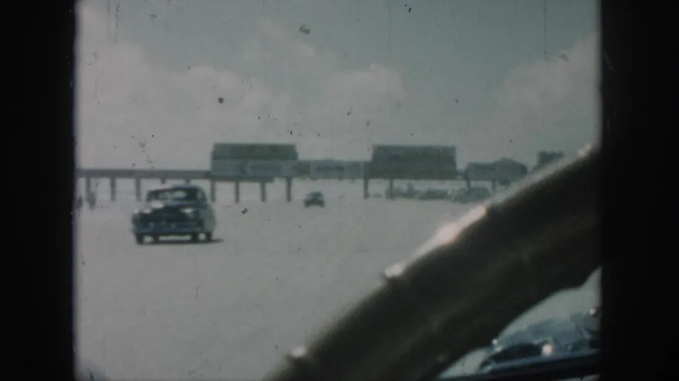 A moving-car view in the New Orleans 1958 clip shows a broad sunlit road, distant vehicles and a long raised structure beyond the windshield.