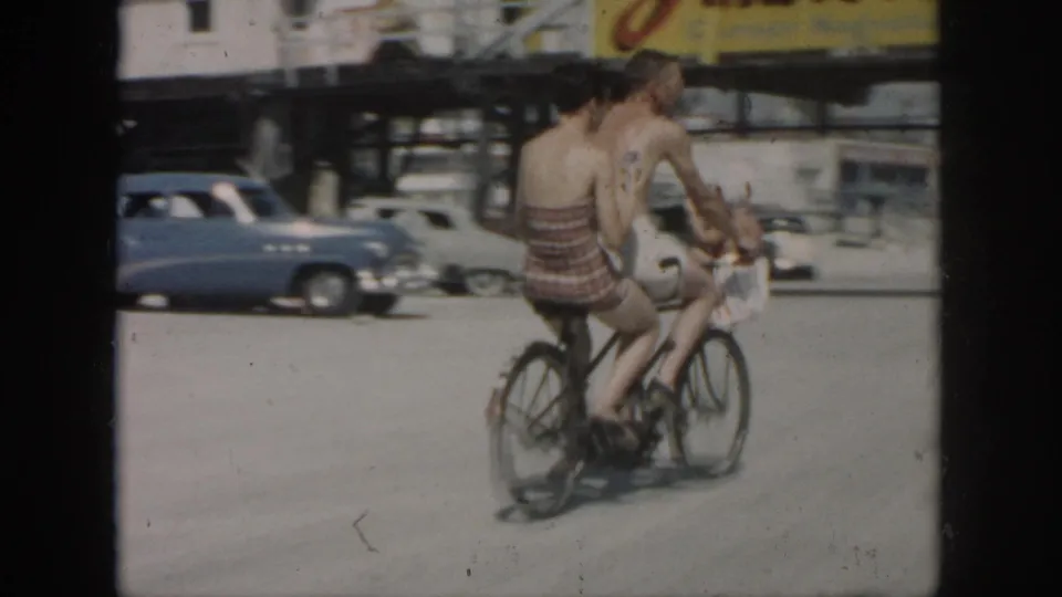 Two riders on a tandem bicycle pass through the 1958 New Orleans road scene, with classic cars and a raised structure in the background.