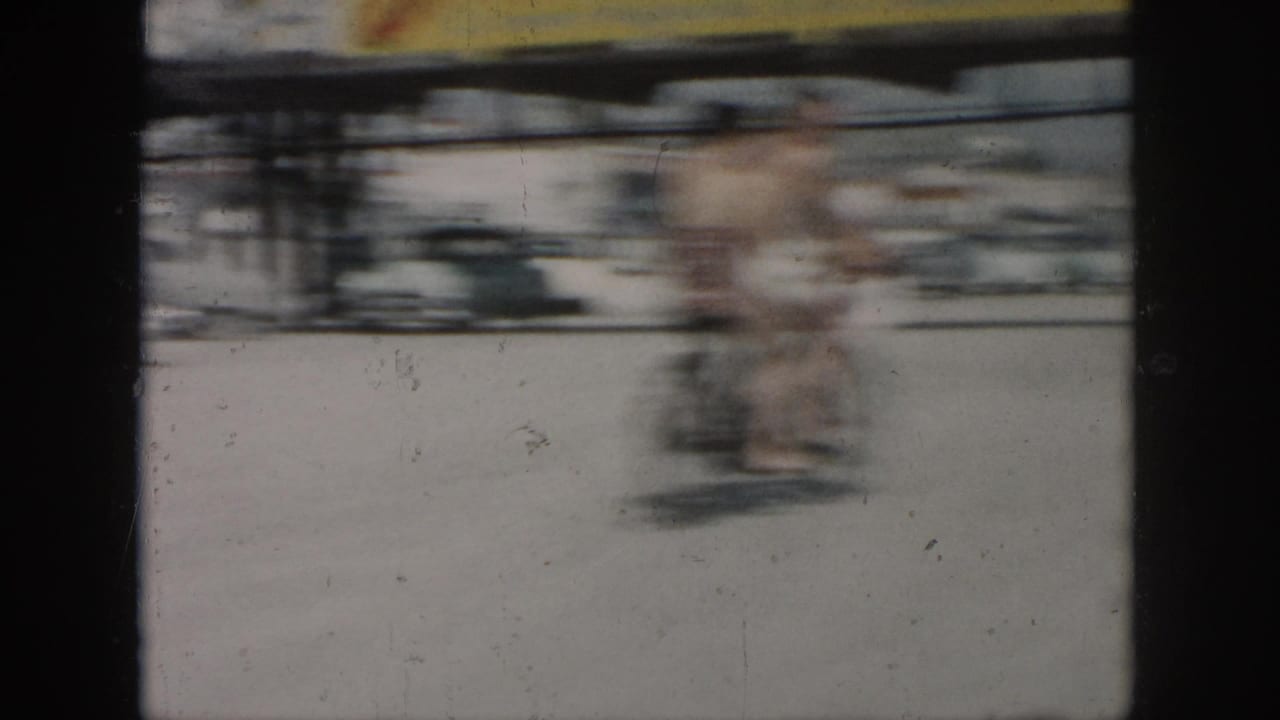 Two men on or near a bicycle pass through a bright outdoor scene in 1958 New Orleans home-movie footage, with motion blur obscuring detail.
