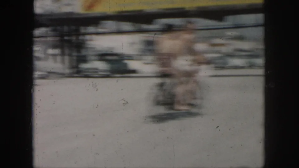 Two men on or near a bicycle pass through a bright outdoor scene in 1958 New Orleans home-movie footage, with motion blur obscuring detail.