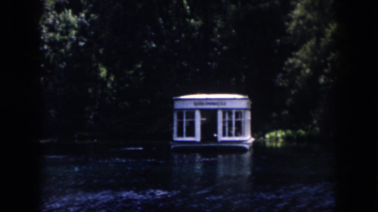 Small white floating structure with windows sits on dark water beside dense trees.