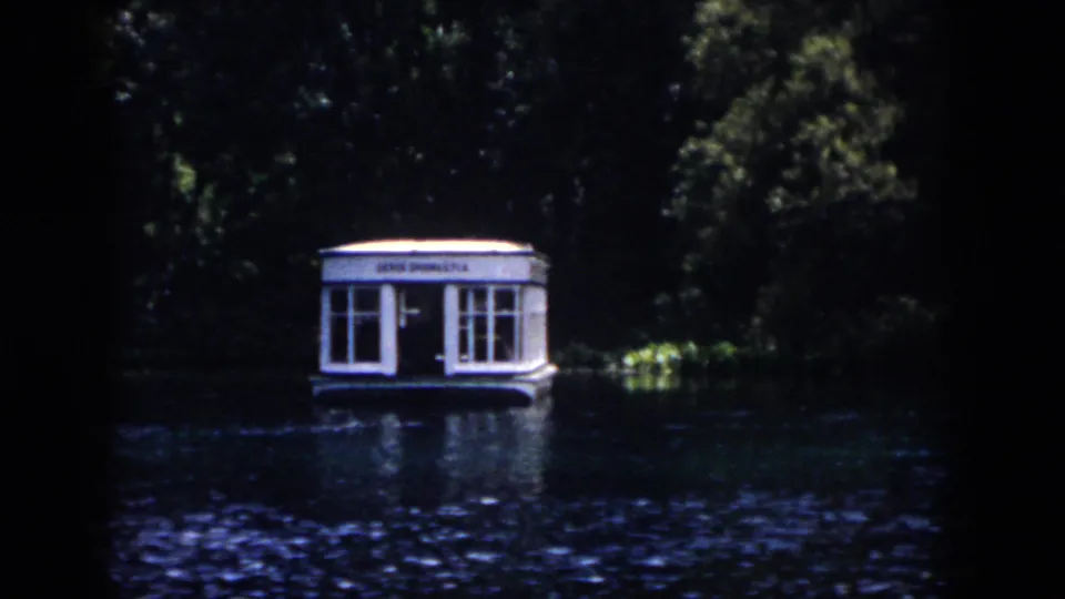 The camera holds on a white floating boathouse or dock structure against dense greenery, with rippled reflections on the water.