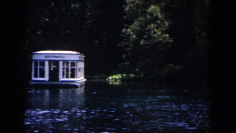 A white floating boathouse or dock structure remains centered across the water, with shaded trees and dark 8mm frame edges around the view.