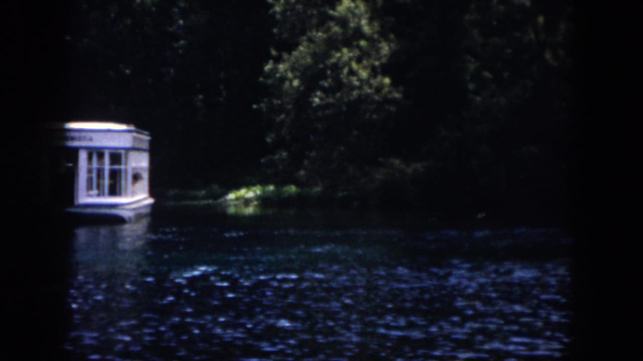 1958 New Orleans home-movie frame showing a small illuminated tour boat moving along dark rippling water near a wooded bank.