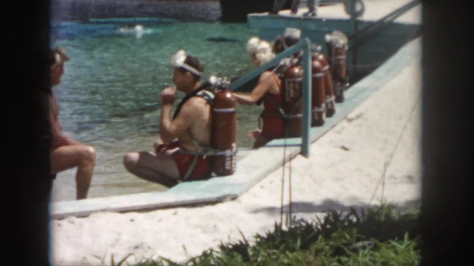 1958 New Orleans dockside frame showing swimmers and divers with large scuba tanks beside clear water.