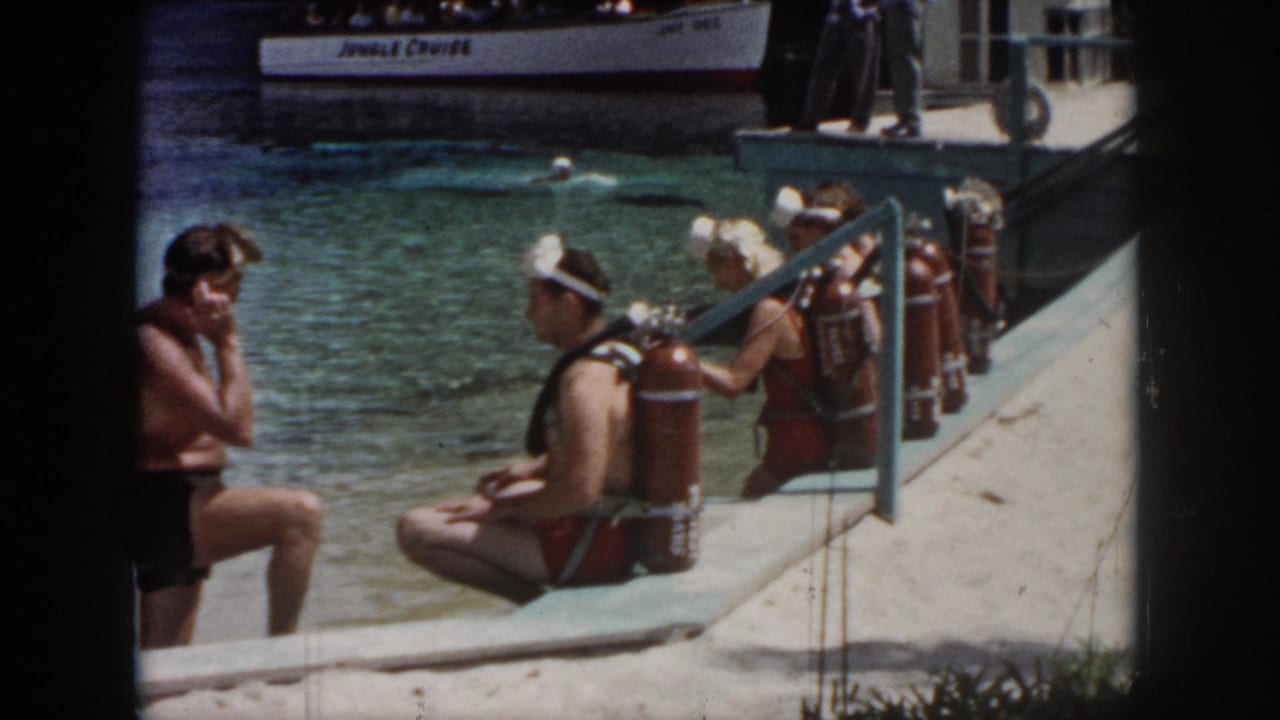 Divers sit by clear water with red tanks while a man in swim trunks adjusts his face near a tour boat.
