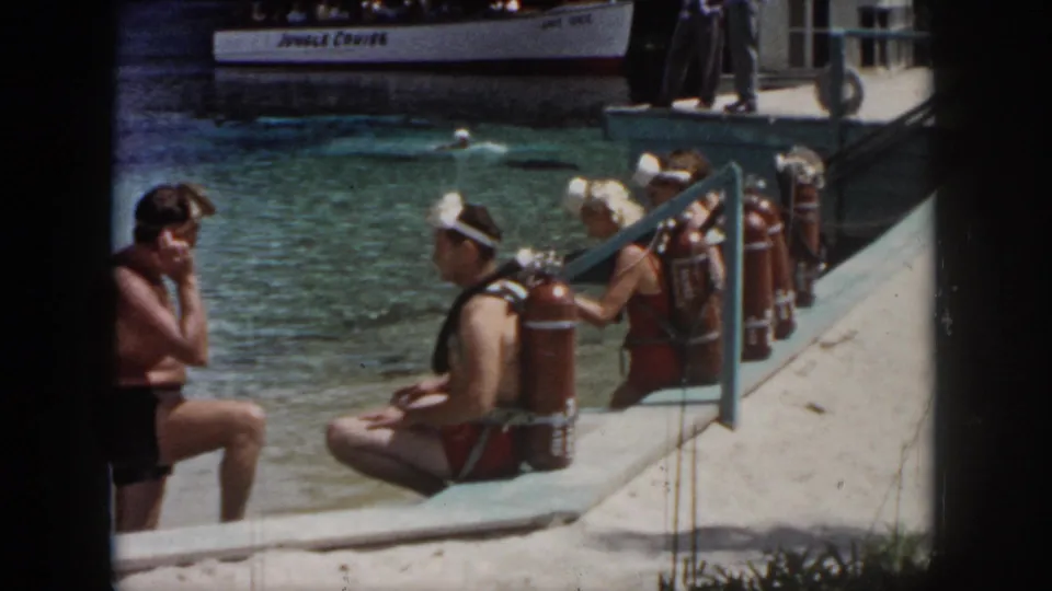 1958 New Orleans home-movie frame of adult divers preparing beside the water, with red tanks, goggles, and a white tour boat behind them.