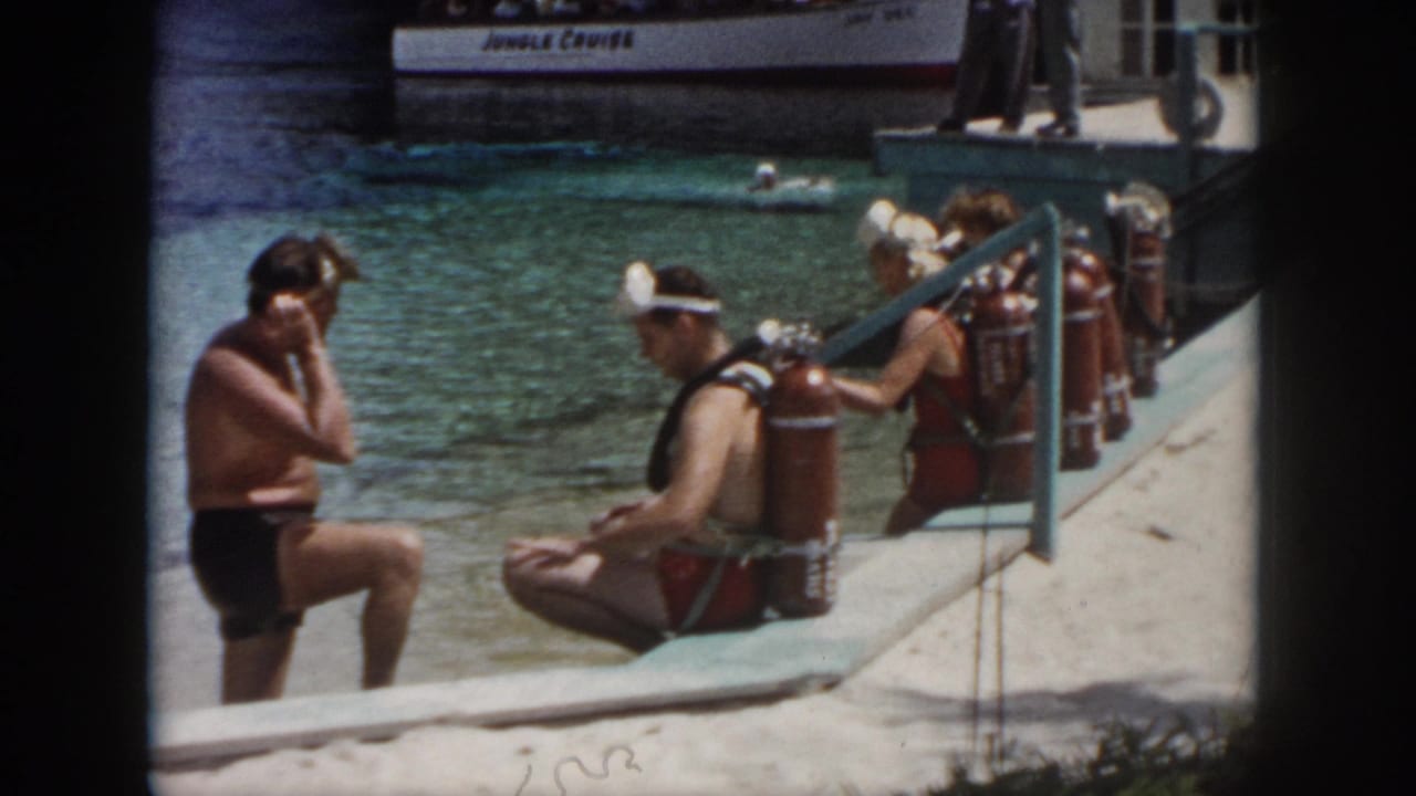 A man stands in shallow water while seated divers with tanks face the water near a docked boat.