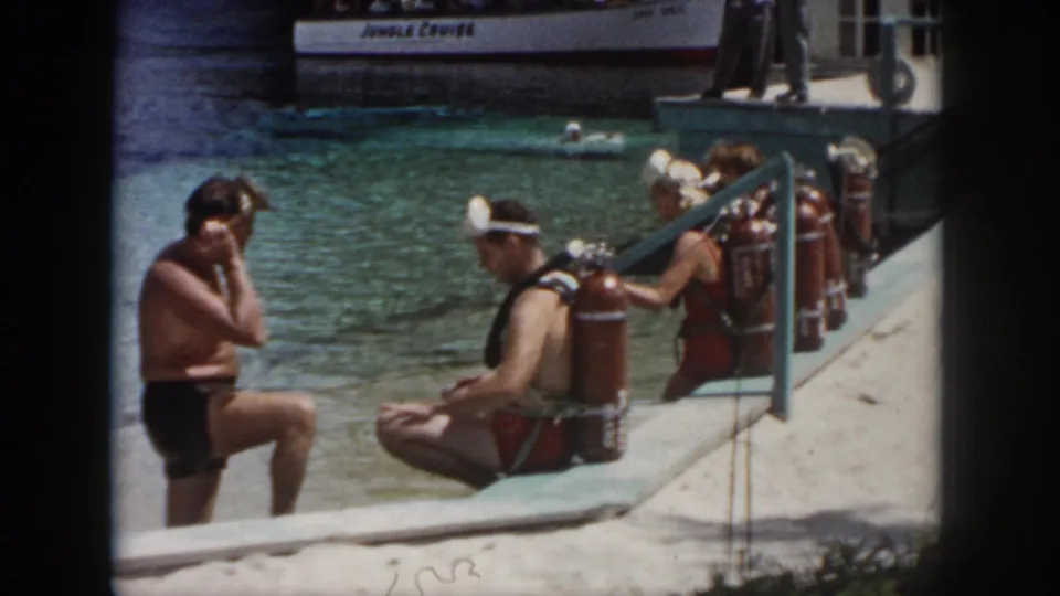 Adult divers with red tanks sit along a waterside ledge in the supplied 1958 New Orleans footage, with a tour boat visible behind them.