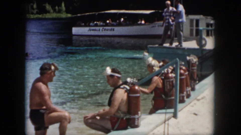 Home-movie frame from the supplied New Orleans 1958 clip showing adult divers with tanks preparing beside clear water and a nearby tour boat.
