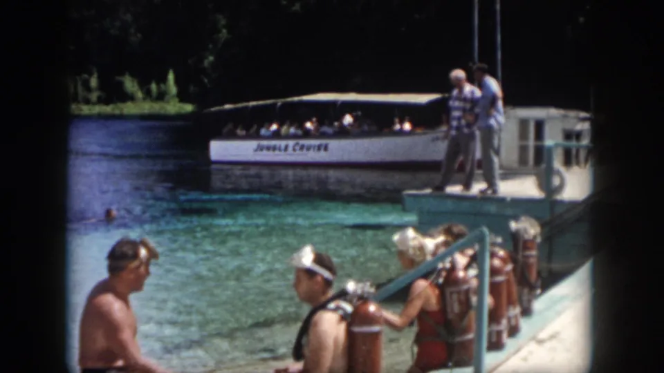 1958 New Orleans home-movie frame of divers in swim gear by the water, with platform figures and a white tour boat in the background.