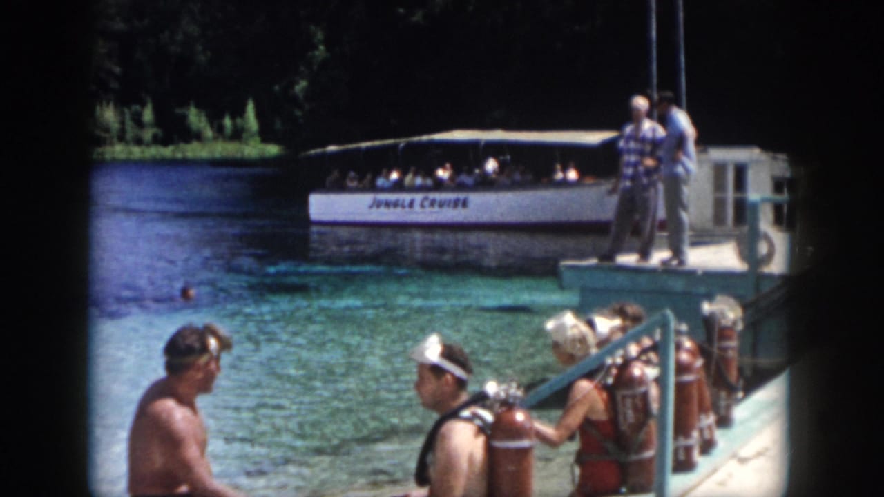 1958 New Orleans home-movie frame showing adults with scuba tanks on a dock near clear water and a Jungle Cruise boat.