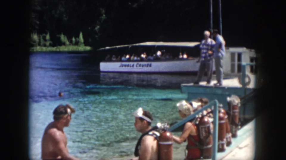 1958 New Orleans home-movie frame showing adults with scuba tanks on a dock near clear water and a Jungle Cruise boat.