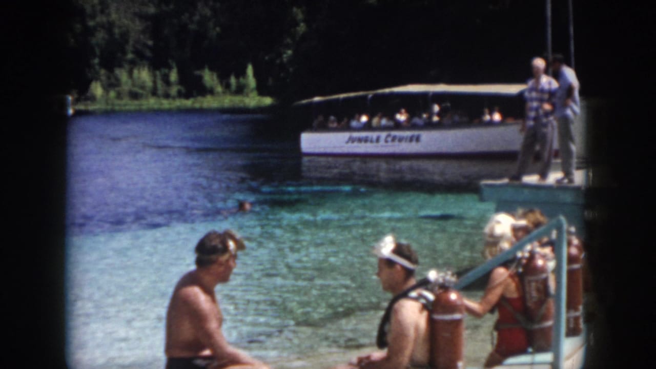 Scuba-equipped adults sit and talk on a dock with a Jungle Cruise boat in the background.