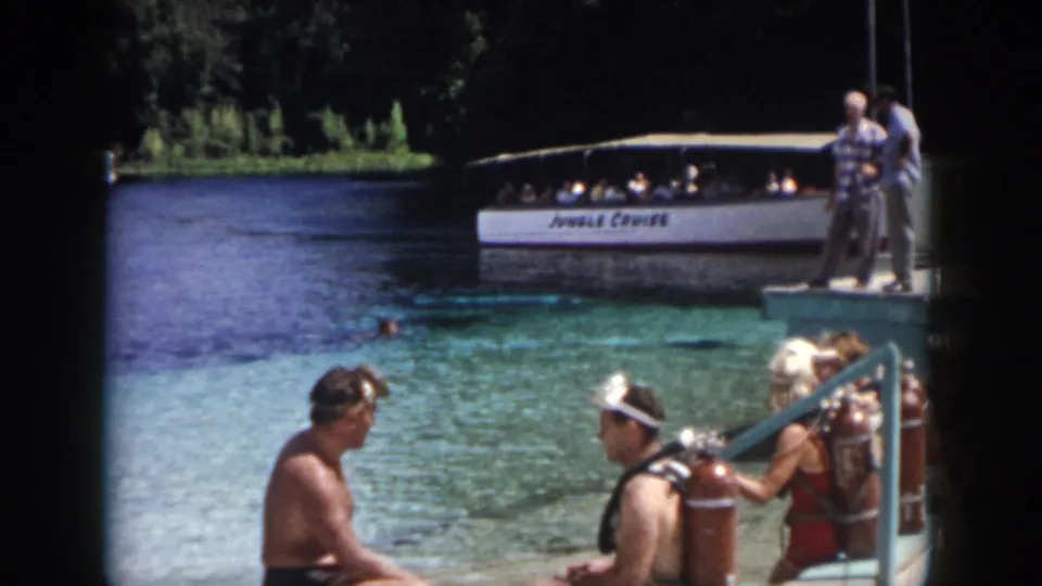 1958 New Orleans footage frame with seated adults wearing vintage scuba gear near a tour boat on a spring-fed waterway.