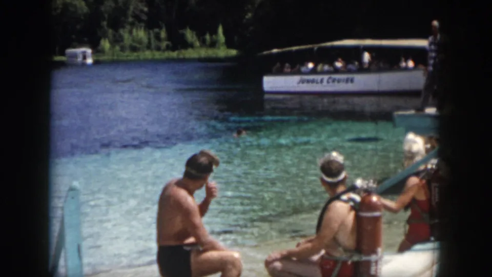 1958 New Orleans home-movie frame showing adults with scuba tanks on a dock, clear water, and a Jungle Cruise boat beyond.