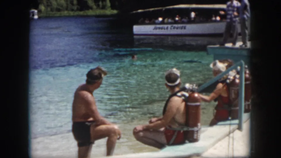 1958 New Orleans frame showing a small group in vintage scuba gear seated by blue-green water as a Jungle Cruise boat passes.